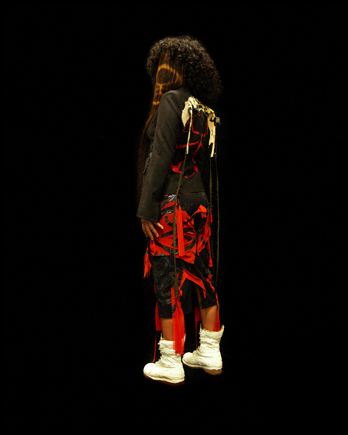 Full body shot of a model facing 3/4 turned away from the camera against a black background. She is wearing a distressed red and black outfit and white boots.